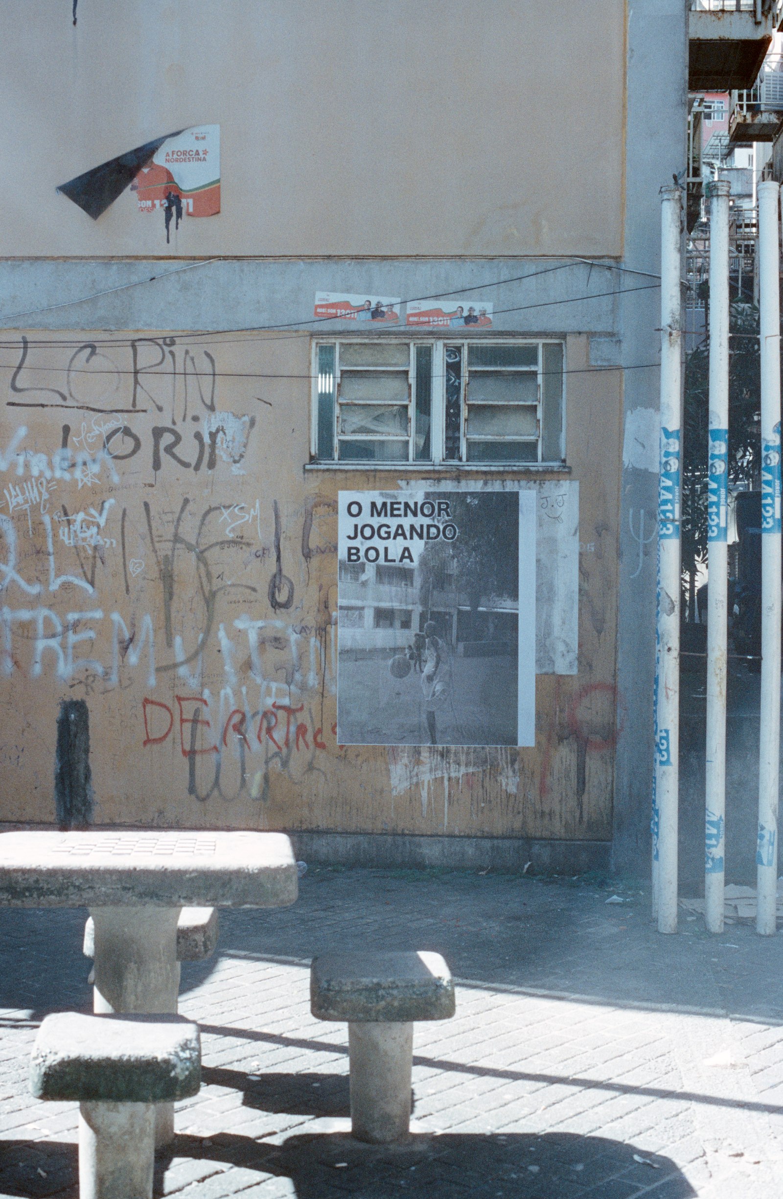 l’affiche « o menor jogando bola » sur un mur, des tables de pierre au premier plan